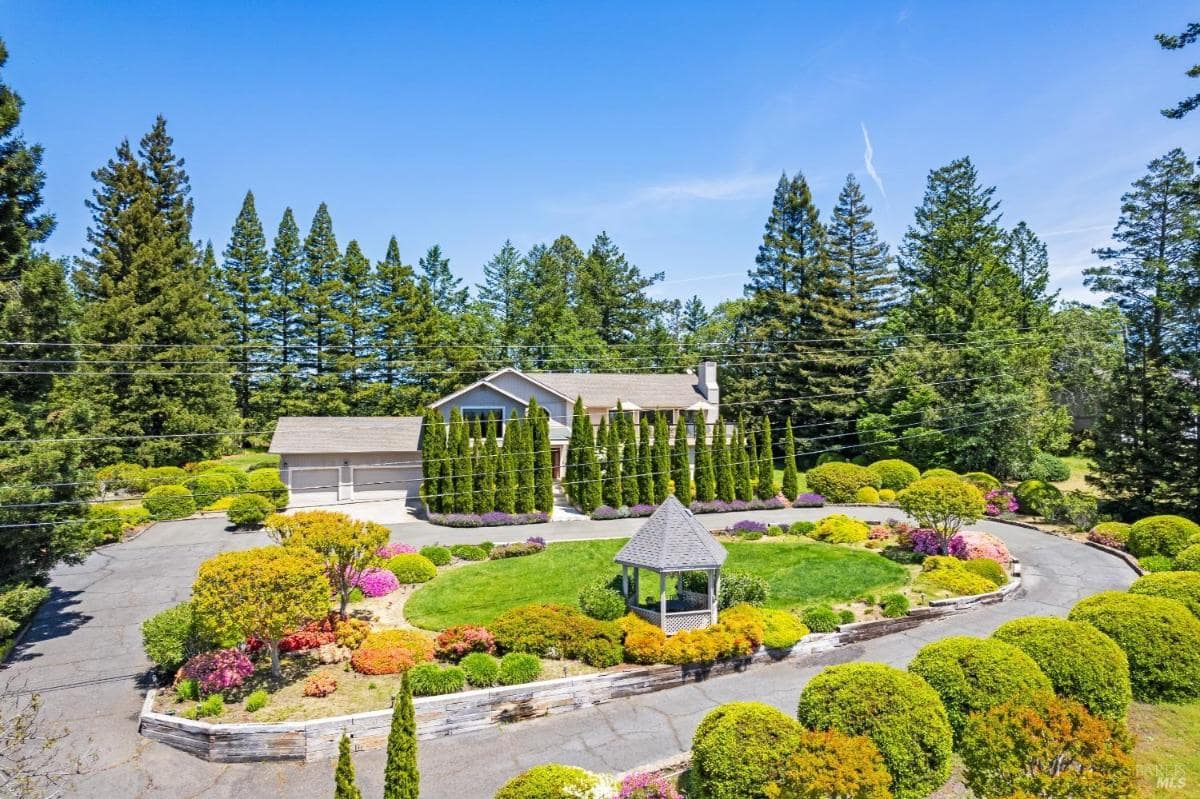 A large house with a circular driveway and a gazebo, surrounded by landscaped gardens and tall trees.