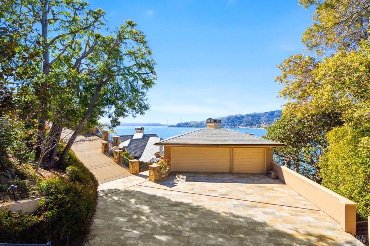 A wide stone driveway leads to a two-car garage, surrounded by trees and overlooking serene bay waters.