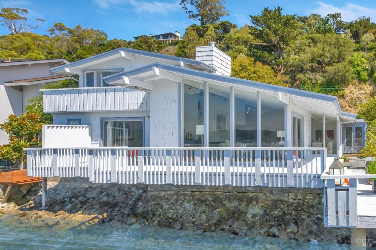 The side exterior view of a white waterfront house with a wraparound deck, situated on a rocky shoreline.