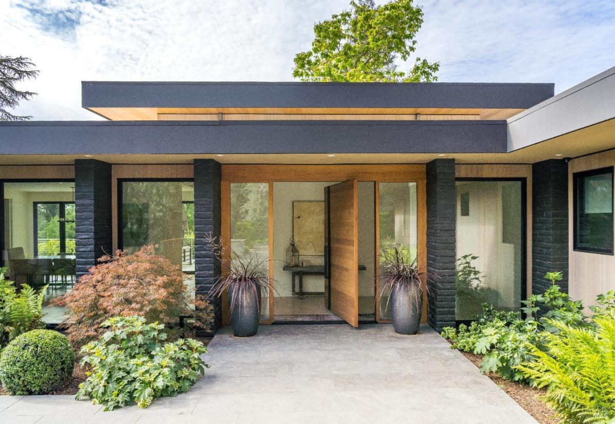 A front exterior view of a house with large windows, a wooden door, and landscaped greenery.