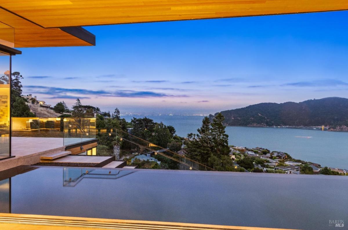 An infinity pool with glass railings and a view of the bay surrounded by hills.