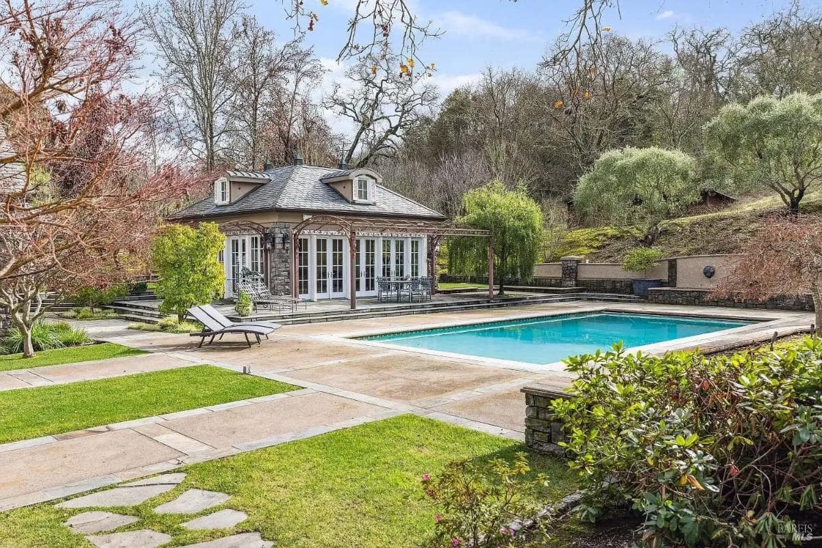 A swimming pool surrounded by landscaped greenery, adjacent to a single-story building with multiple French doors and outdoor seating areas.