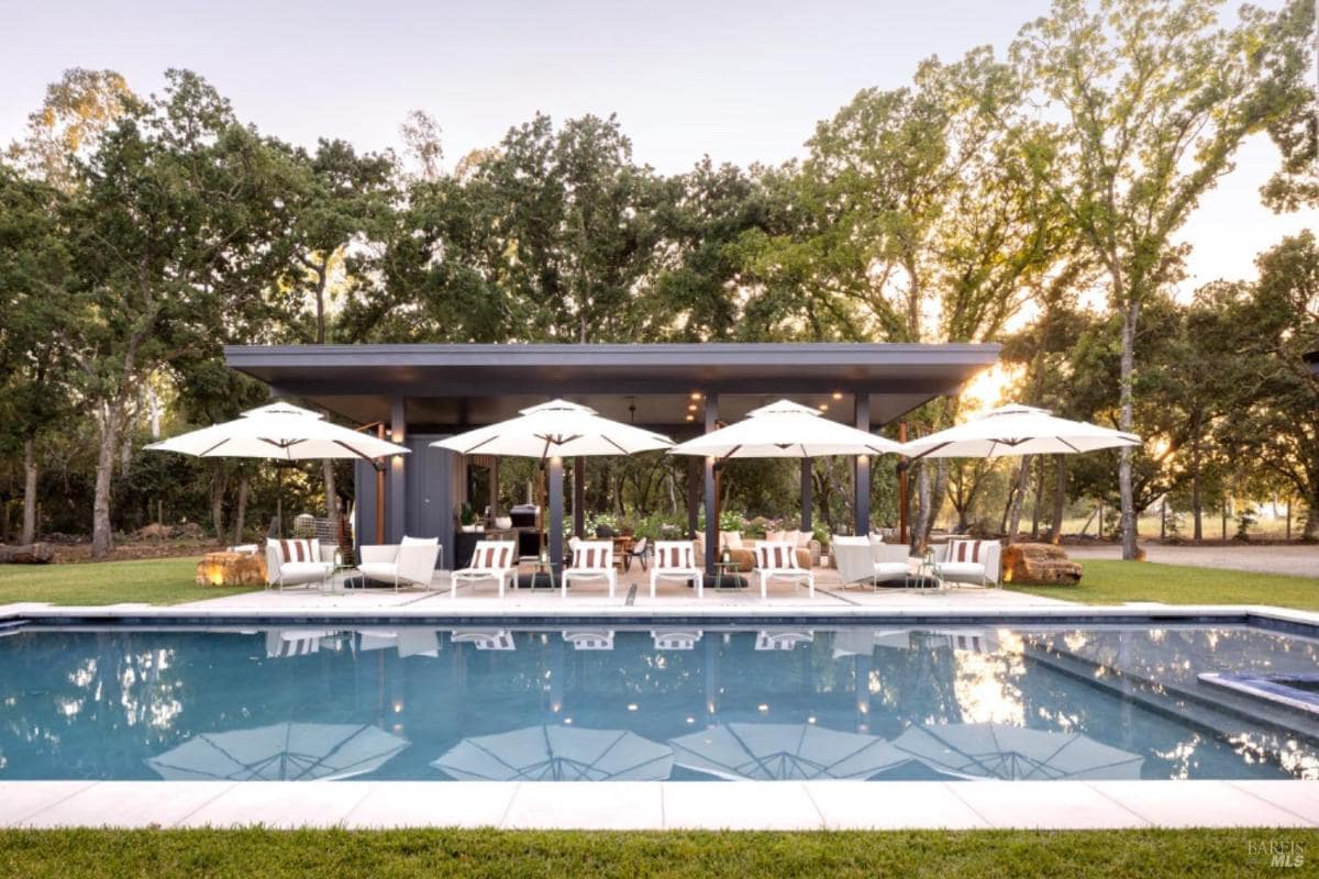 A pool with surrounding lounge chairs, umbrellas, and a covered outdoor living area set against a backdrop of tall trees.