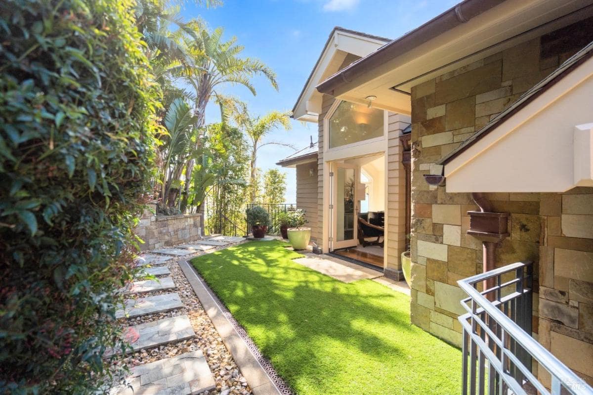 A landscaped garden with a stone path, bordered by lush greenery and palm trees. The home's exterior features stone accents and large glass doors.