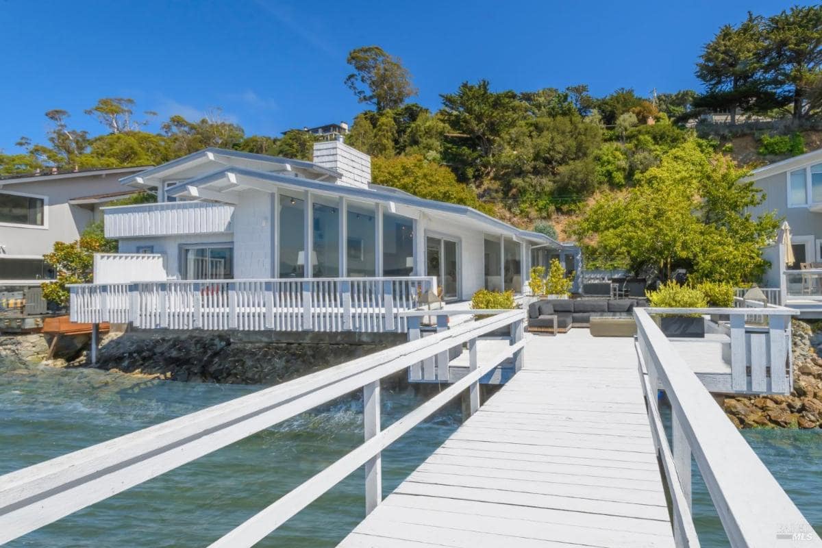 A waterfront house with a wooden deck leading to a dock, showcasing outdoor seating areas and large windows.