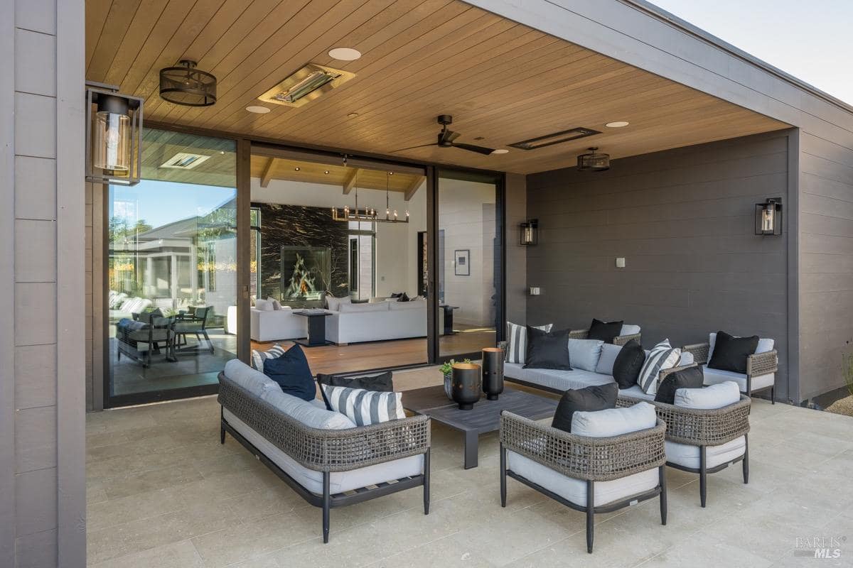 A rear patio with sofas and chairs, under a covered patio with ceiling heaters and fans, adjacent to sliding glass doors.