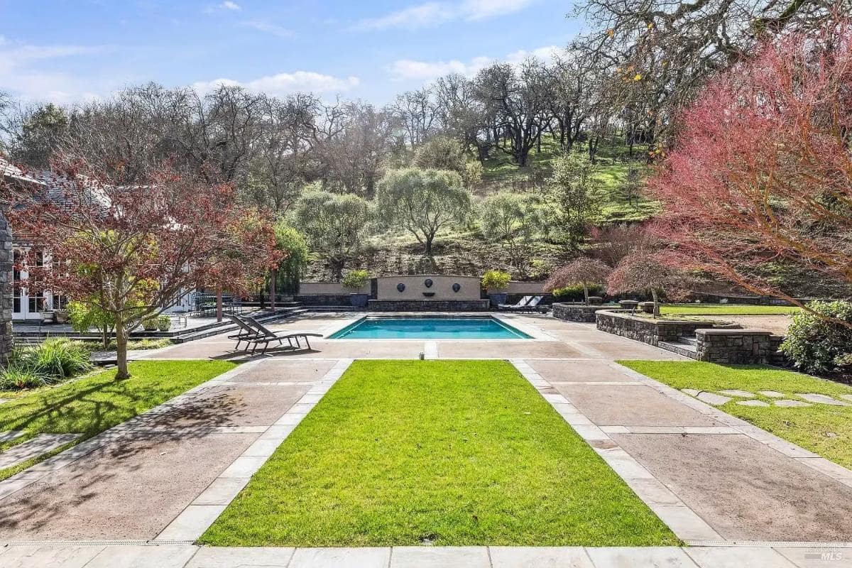 A centered view of a swimming pool with green lawns on both sides and a retaining wall with decorative water features in the background.