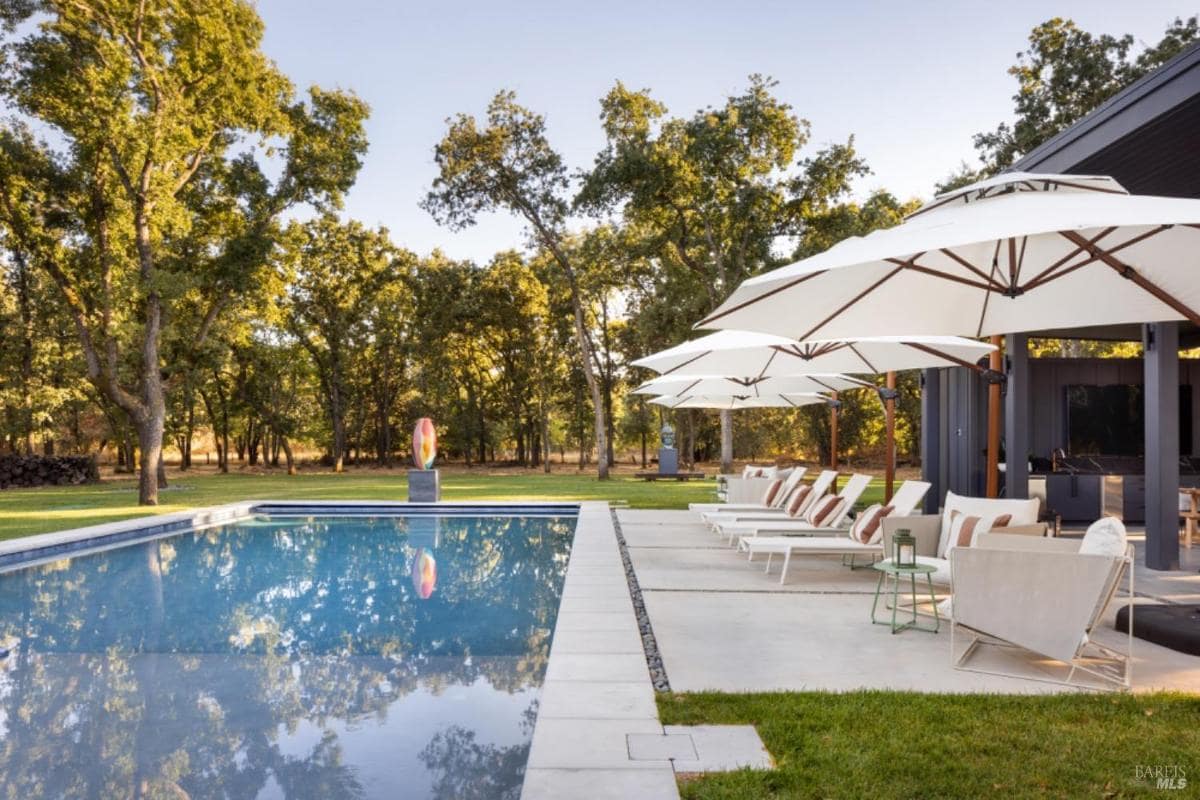 A poolside area with lounge chairs, large umbrellas, and a sculpture surrounded by trees.