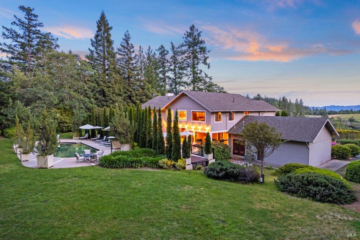 A picturesque view of a large house with a pool, surrounded by lush greenery and tall trees, set against a beautiful sunset sky.