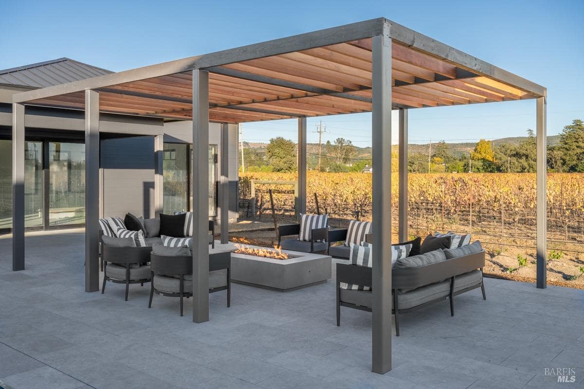 An open pergola with a fire pit, surrounded by cushioned seating, overlooks a vineyard.