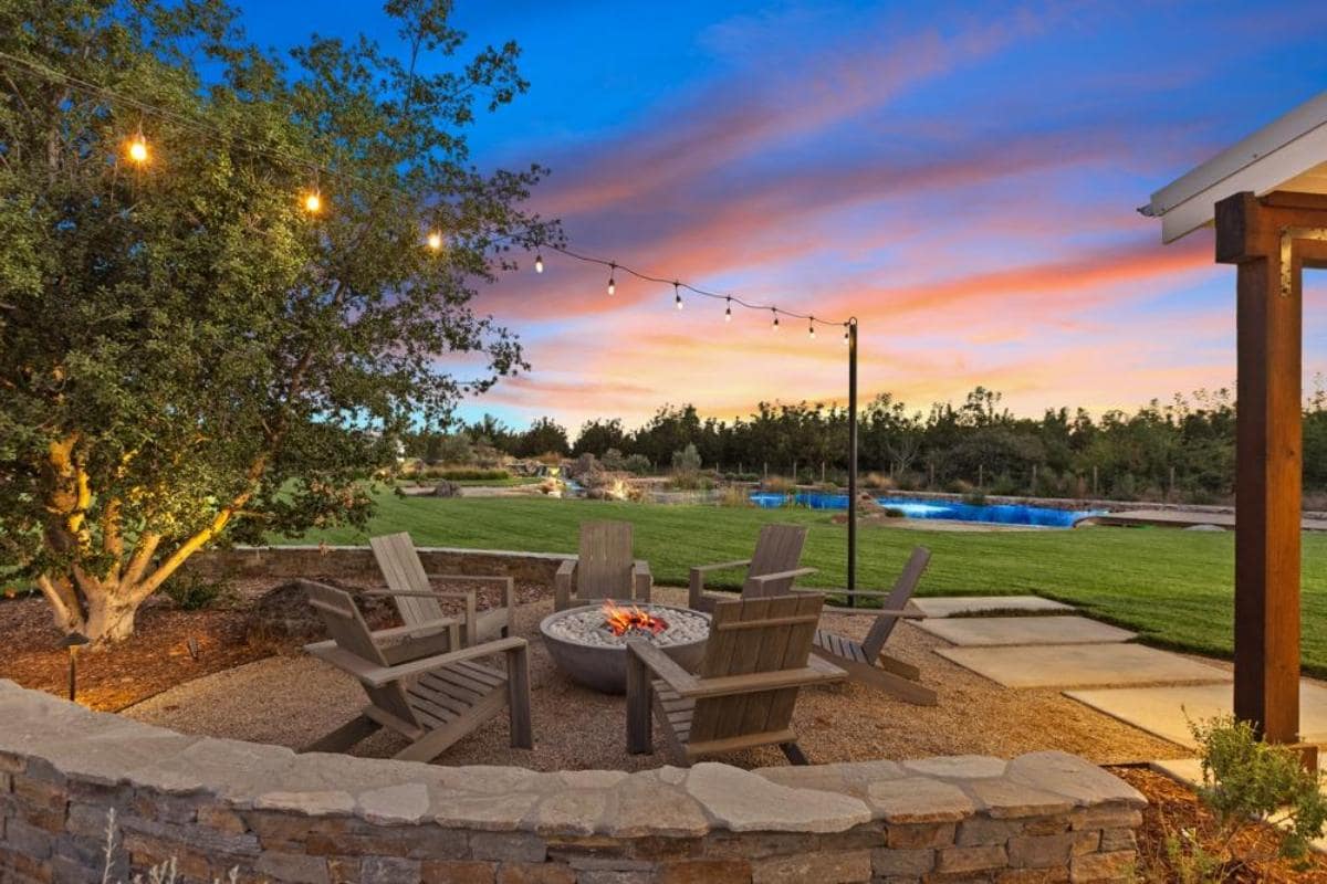 A cozy fire pit area with Adirondack chairs, surrounded by greenery and string lights, set against a colorful sunset and a swimming pool in the background.