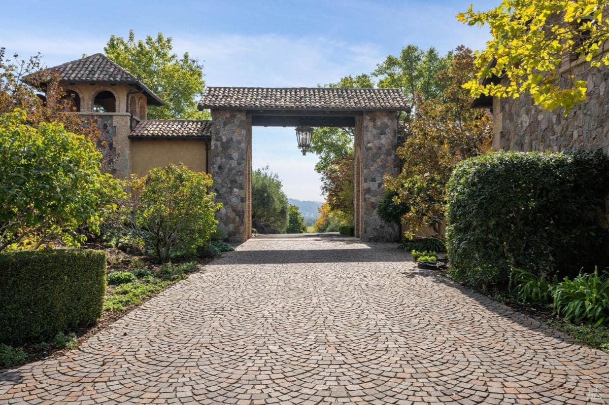 A cobblestone driveway leading through a stone archway with a tiled roof, flanked by trees and shrubs.