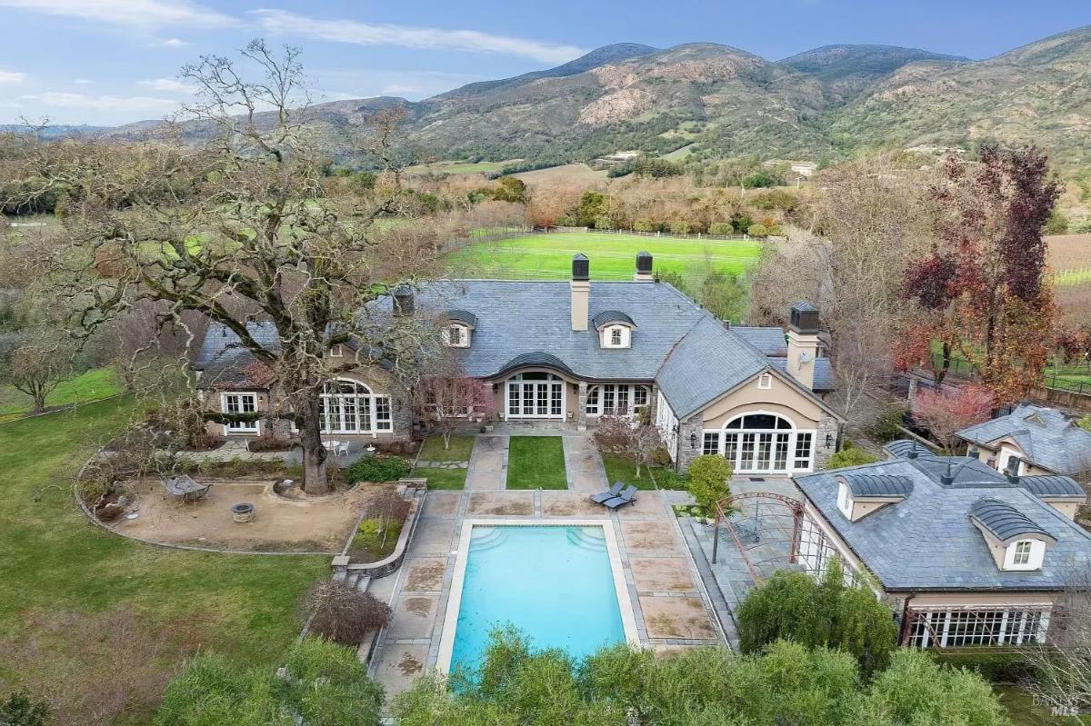 An aerial view of a mansion featuring a swimming pool, main residence, surrounding greenery, and nearby hills