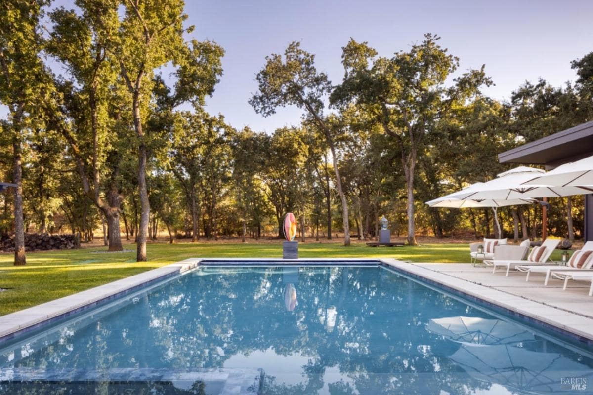 A rectangular pool with reflections of trees and umbrellas, surrounded by lounge chairs and greenery.