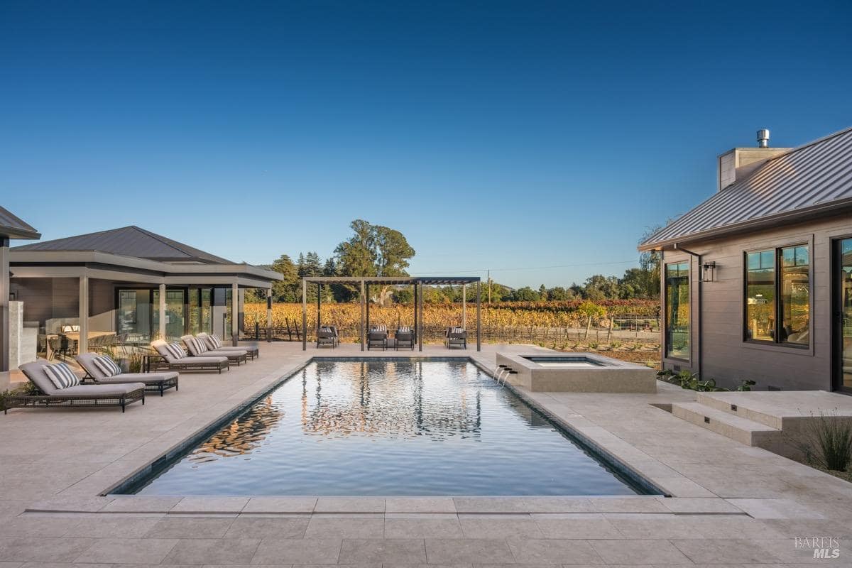 A swimming pool with surrounding lounge chairs and a pergola overlooks a vineyard.