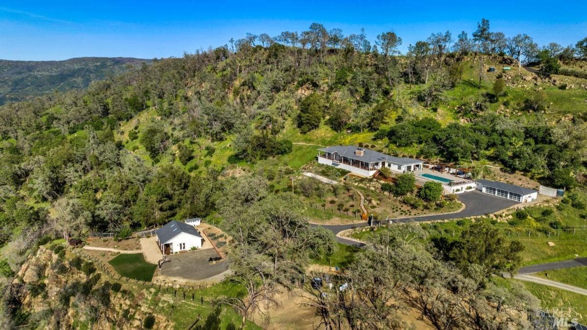 A property on a hillside featuring multiple buildings, a pool, and a winding road surrounded by greenery.