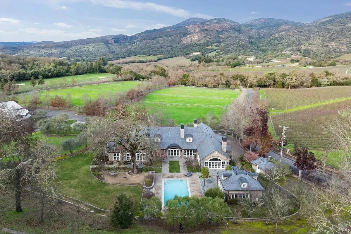 Aerial view of a large mansion showcasing a residence, swimming pool, landscaped gardens, and surrounding farmland with hills in the background.