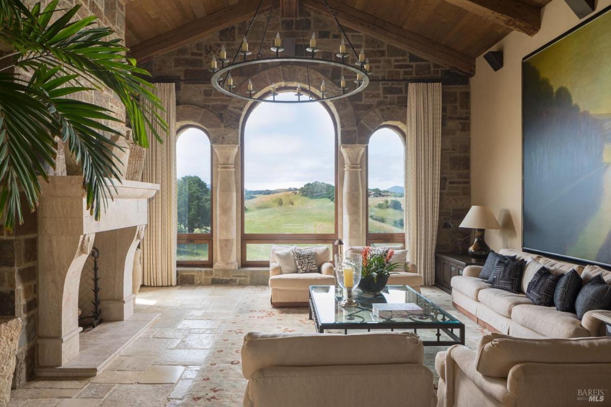 A living room with a stone fireplace, large windows overlooking greenery, and a coffee table with flowers.