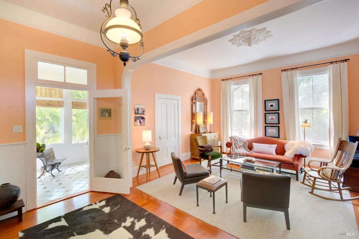 A living room with peach walls, large windows with curtains, various chairs, a glass coffee table, and decorative items.