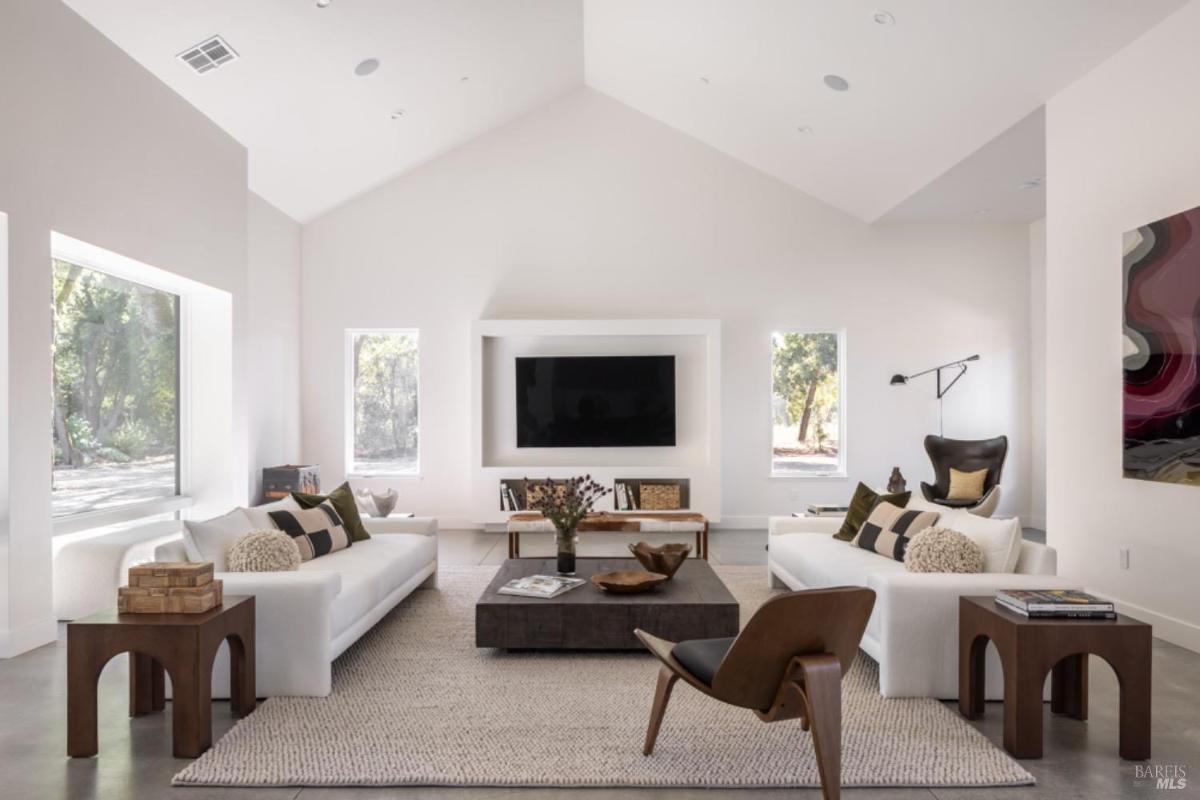 A living room with vaulted ceilings, large windows, and a centered TV wall, featuring two facing sofas, a rug, and modern furniture.