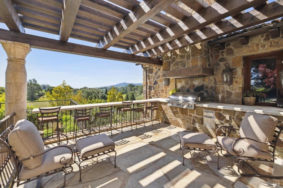 An outdoor kitchen with a stone grill, bar stools, and comfortable seating under a wooden pergola.