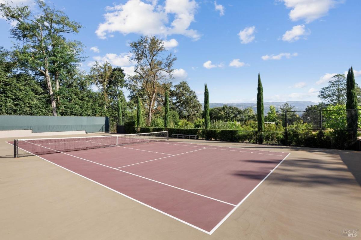 A tennis court surrounded by tall trees and a backdrop of hills under a clear sky.