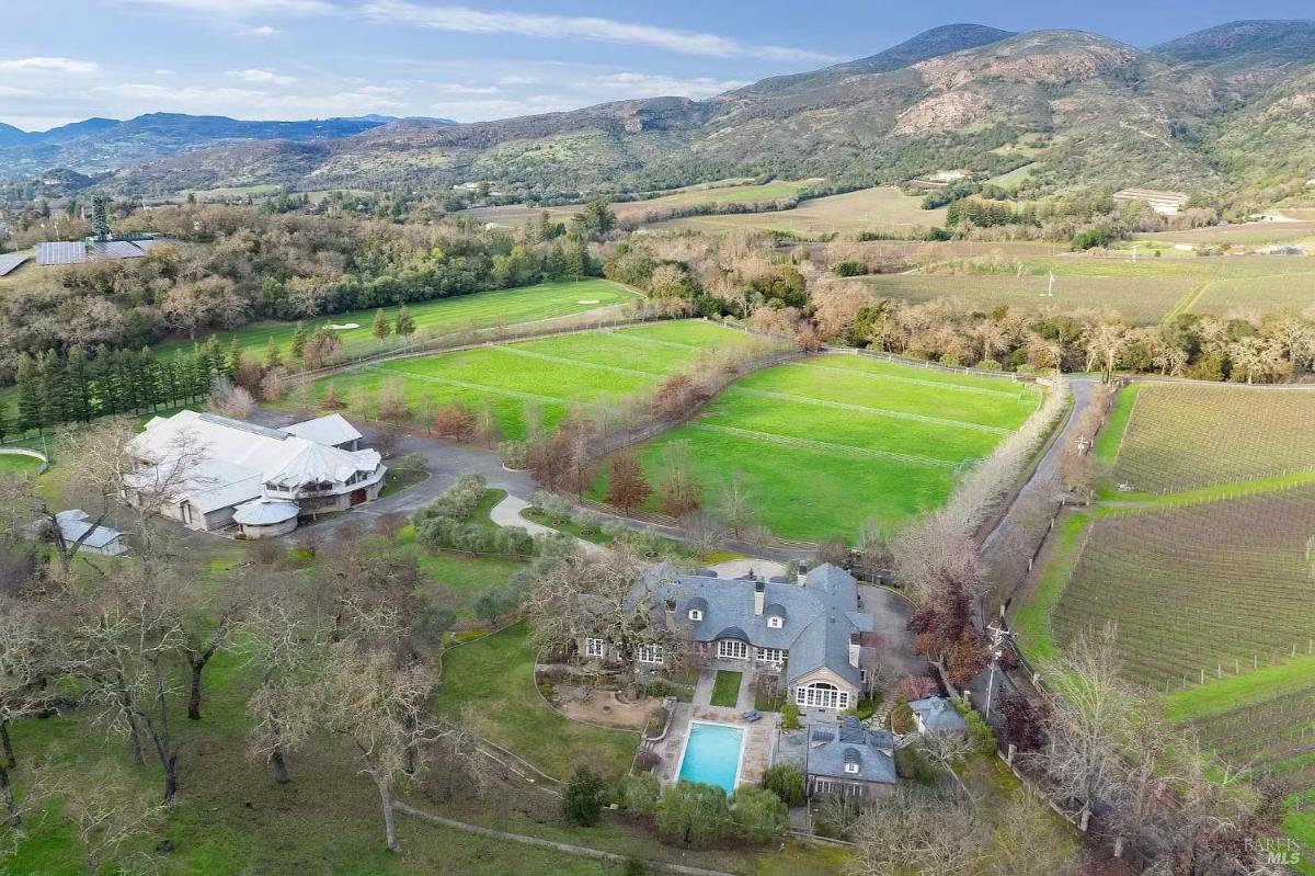 Wide aerial view of a sprawling property including a residence, swimming pool, large barn-like structure, landscaped grounds, and adjacent fields and hills.