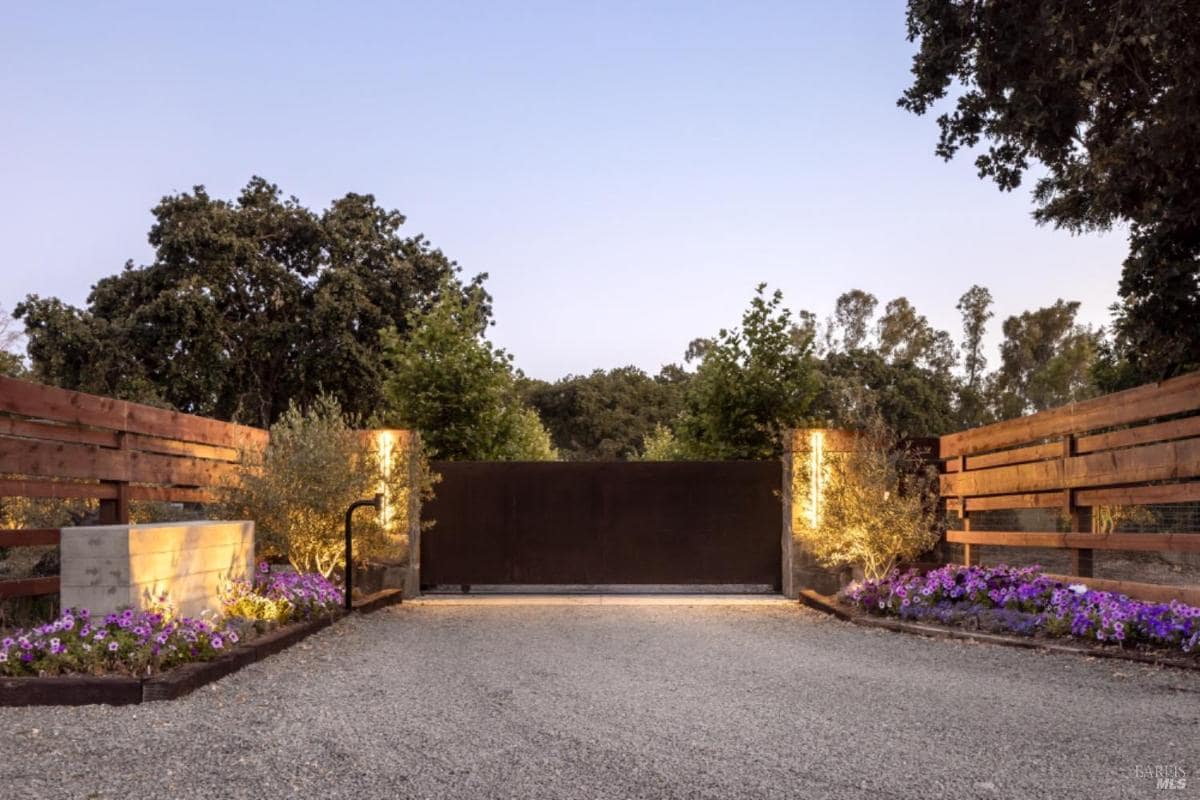 A gated entrance with wooden fencing, illuminated pillars, and landscaped flower beds.