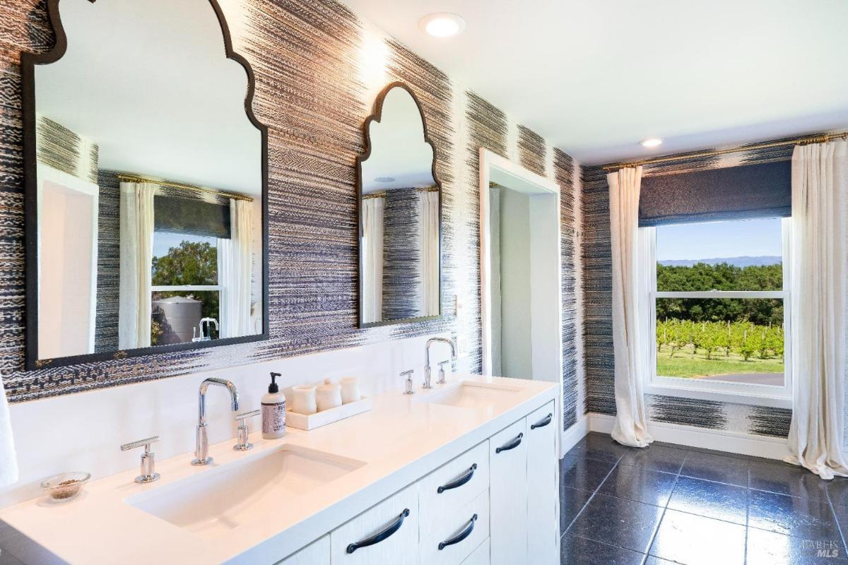 A modern bathroom with double sinks, large mirrors, and a window overlooking a vineyard. 