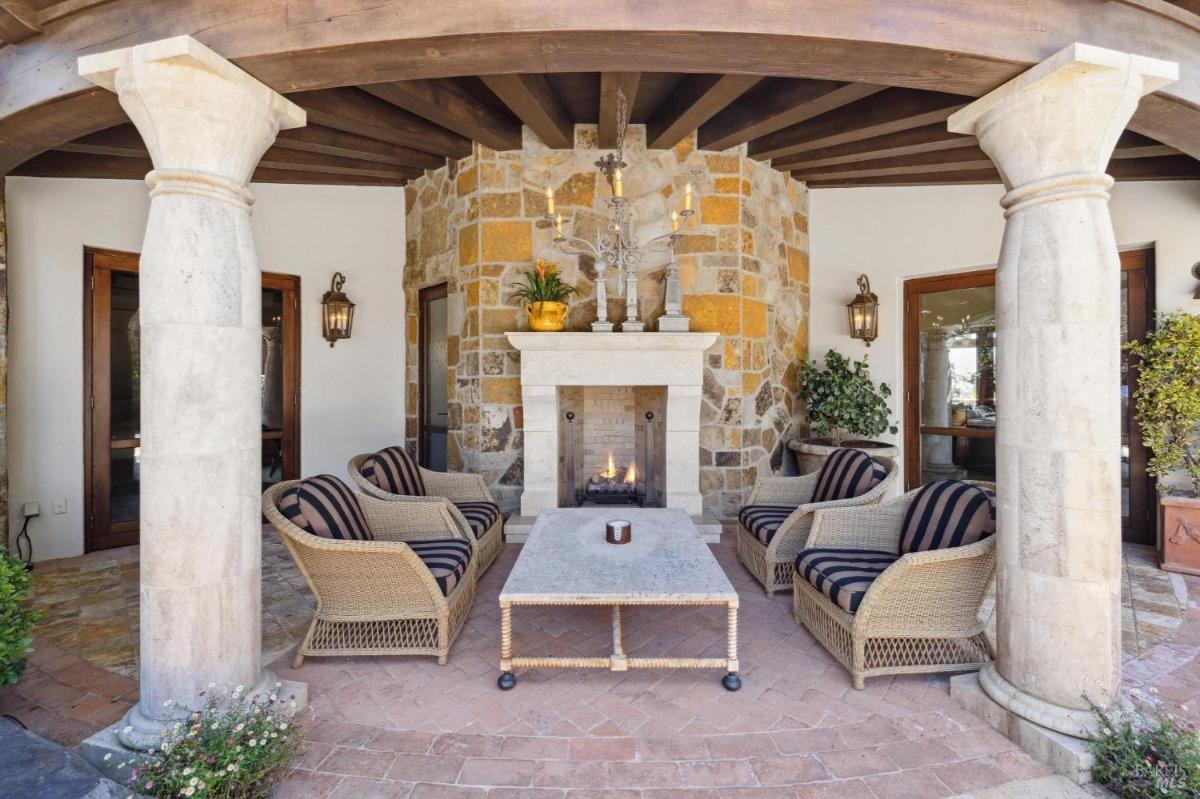 A patio featuring a stone fireplace, surrounded by columns and comfortable chairs with a central table. 