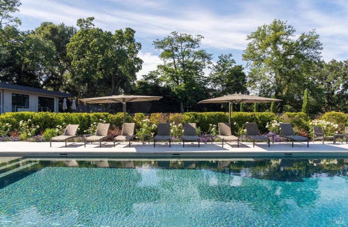 A poolside view with lounge chairs, umbrellas, and a landscaped background of hedges and trees.