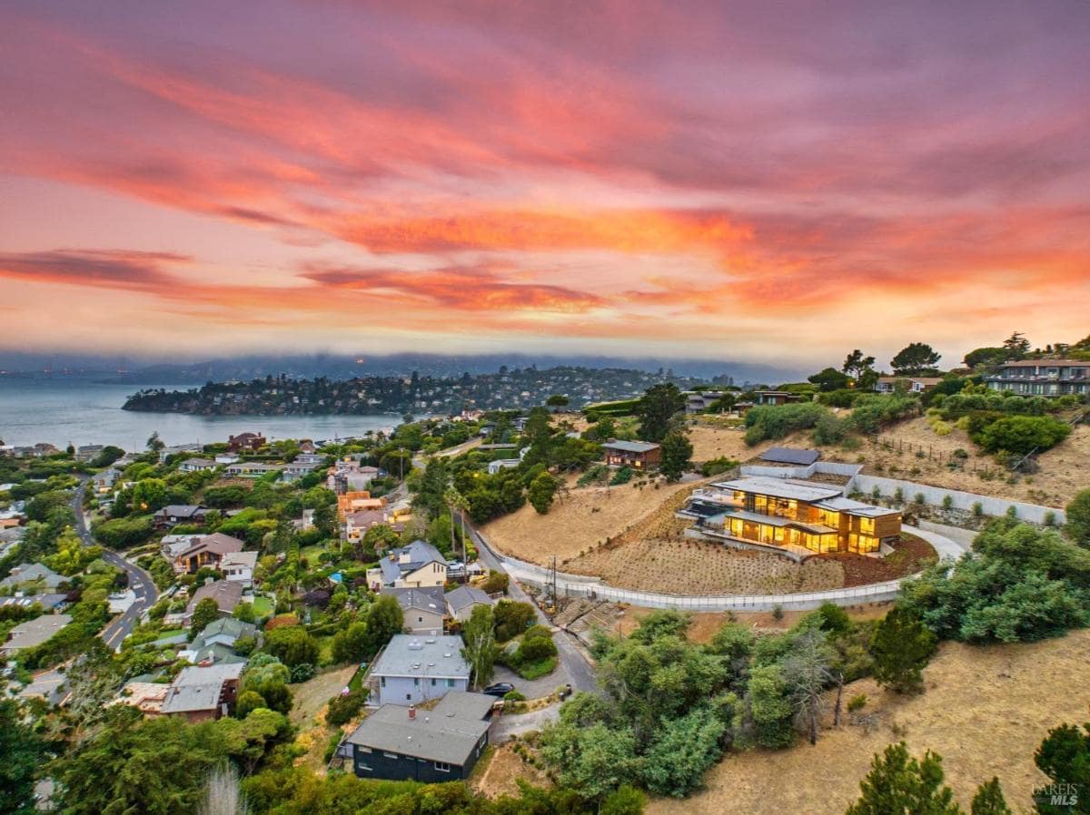 A large hillside home with modern architecture, surrounded by greenery, overlooking a coastal neighborhood.