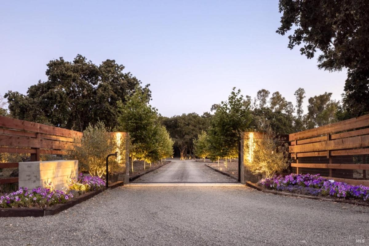 An illuminated driveway entrance with wooden fencing, landscaped flower beds, and rows of trees lining a gravel path.