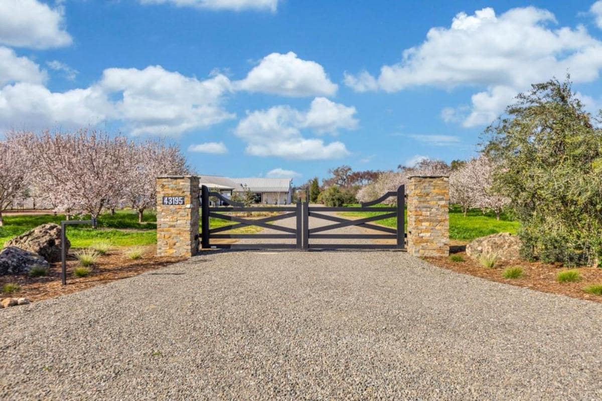 Black gate at the entrance of a gravel driveway lined with trees and rocks.