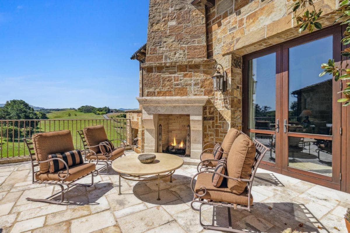 An outdoor seating area with a stone fireplace, featuring comfortable chairs arranged around a circular table. 