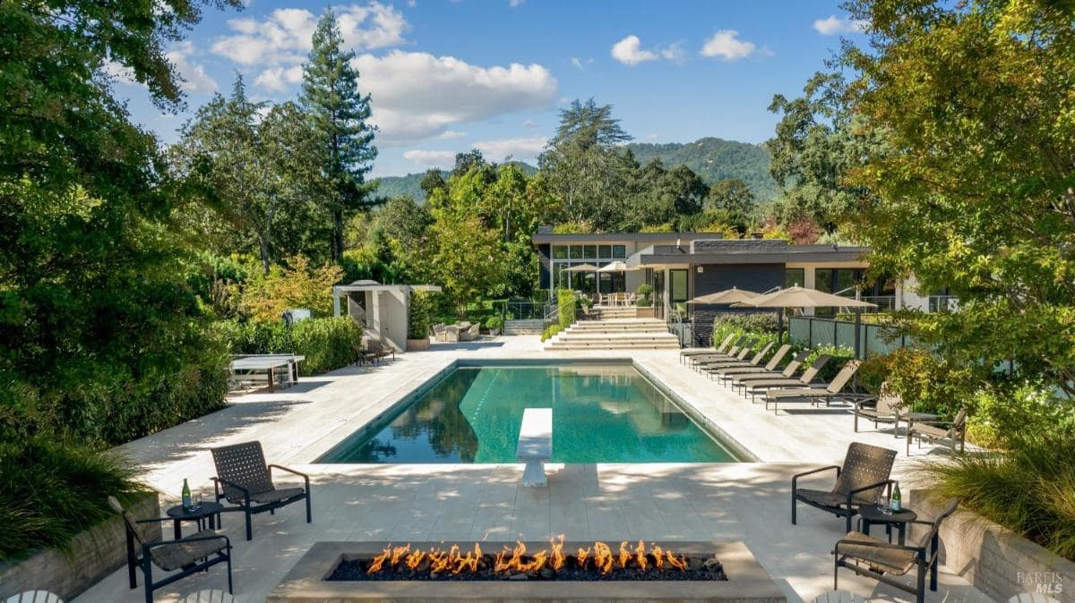 A rectangular pool with a fire pit in the foreground and a modern house in the background surrounded by trees.