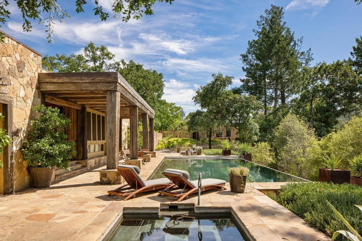 Stone patio with lounge chairs next to an infinity pool surrounded by greenery.