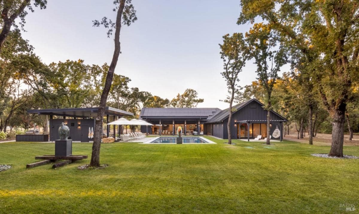 A landscaped backyard featuring a pool, outdoor pavilion, and modern black home surrounded by tall trees and open green lawn.