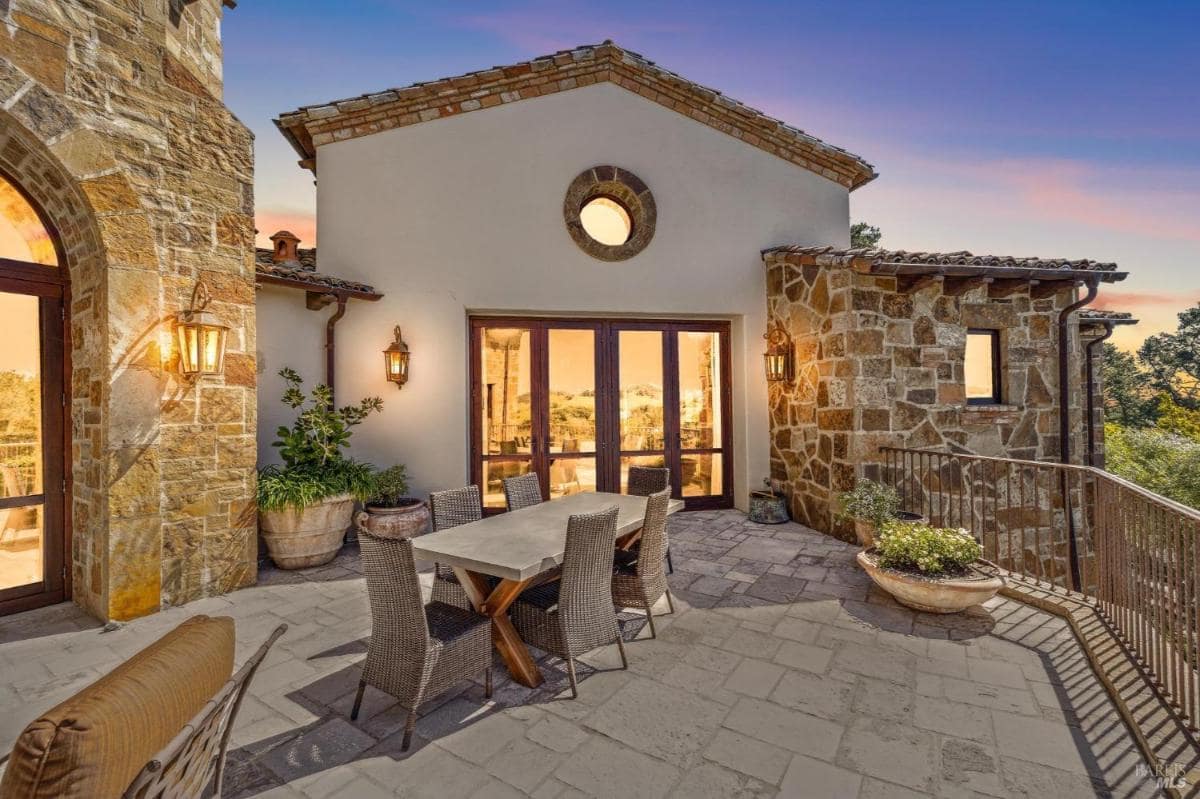 An outdoor dining area with a long table surrounded by wicker chairs, set against a backdrop of a beautifully designed stone and stucco building. 