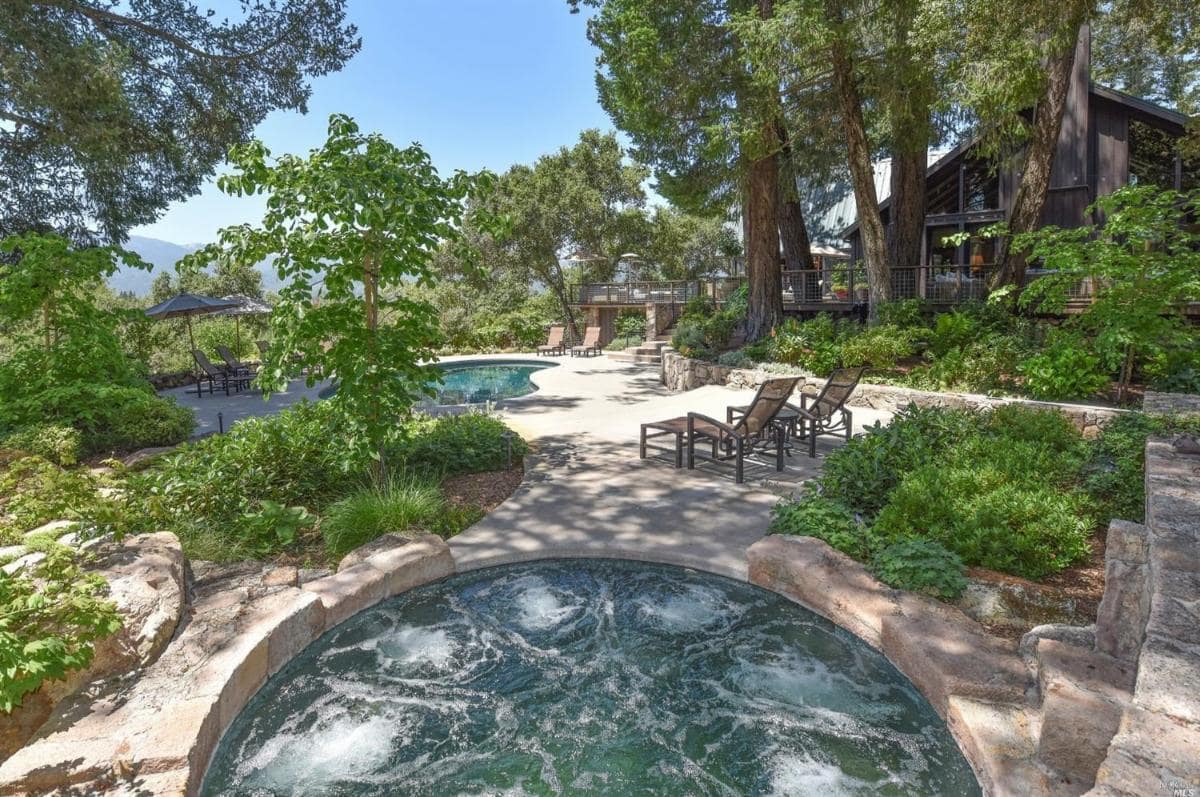 Pool area with surrounding lounge chairs and a hot tub under trees.