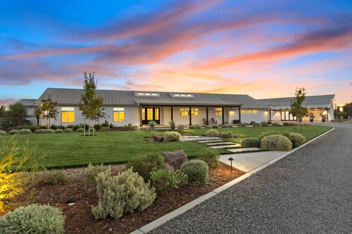 Modern house with a metal roof, surrounded by landscaped gardens and a gravel pathway, set against a colorful sunset sky.