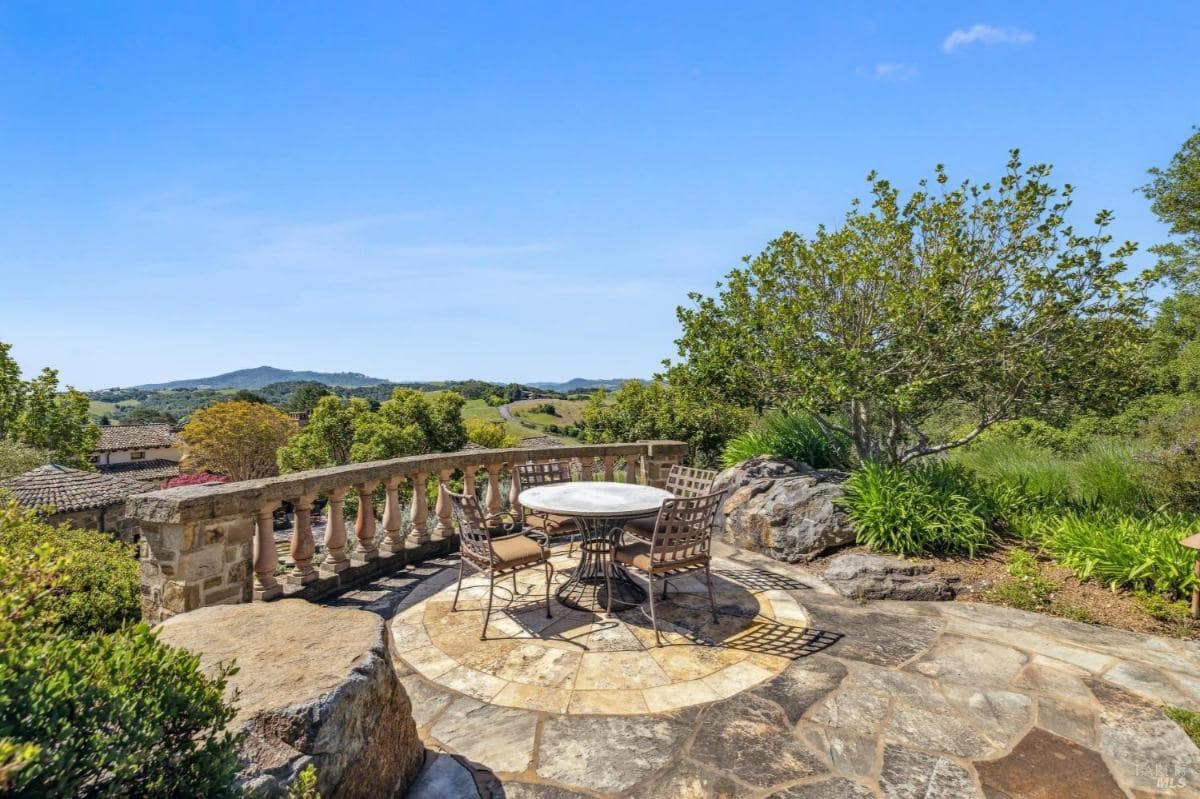 Outdoor seating area with a stone patio, featuring a round table and chairs, surrounded by greenery and a scenic view of hills and trees in the background.