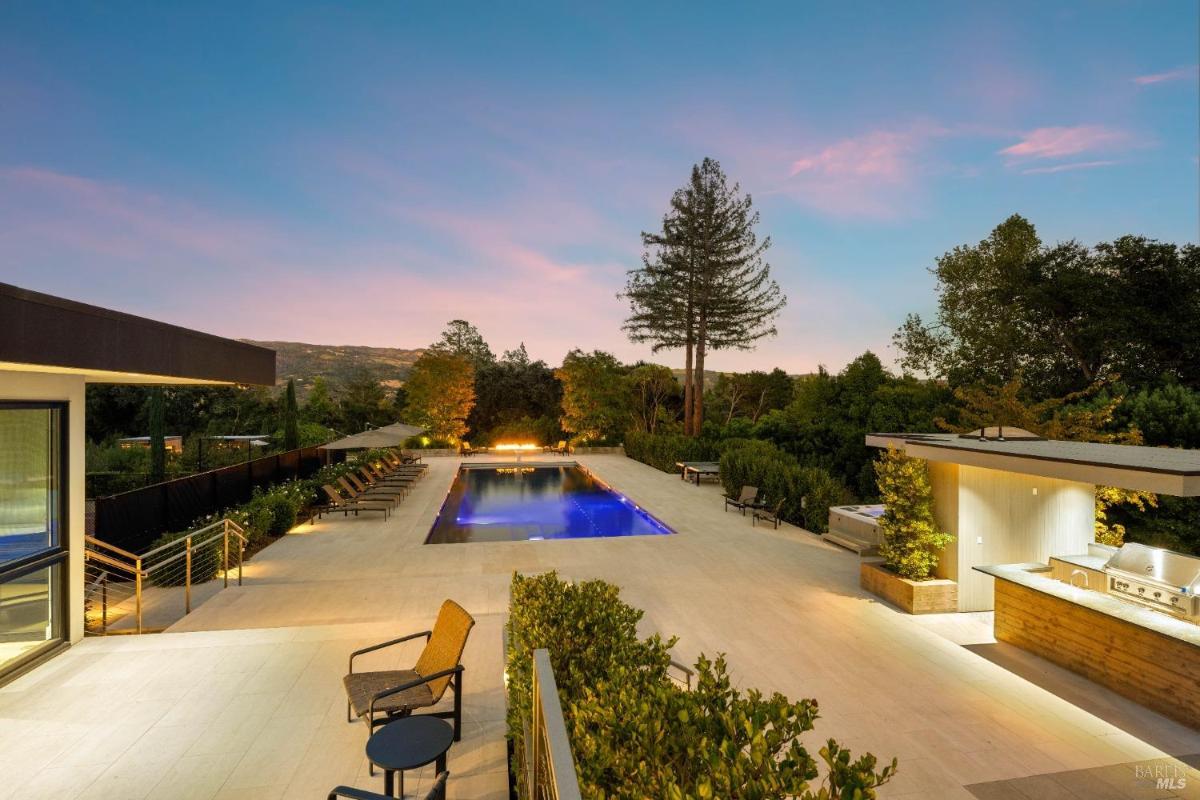 A rectangular swimming pool illuminated in the evening, surrounded by lounge chairs, a fire pit at one end, and an outdoor kitchen to the side.