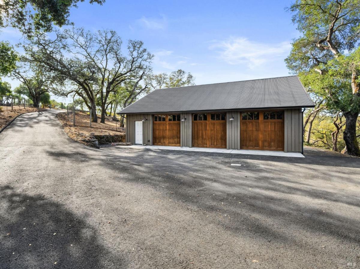 Driveway leading to a detached garage with a metal roof and wooden doors, surrounded by trees.