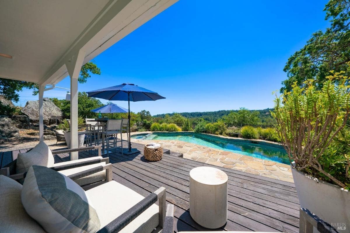 A patio area with seating, a table, an umbrella, and a swimming pool in the background. 