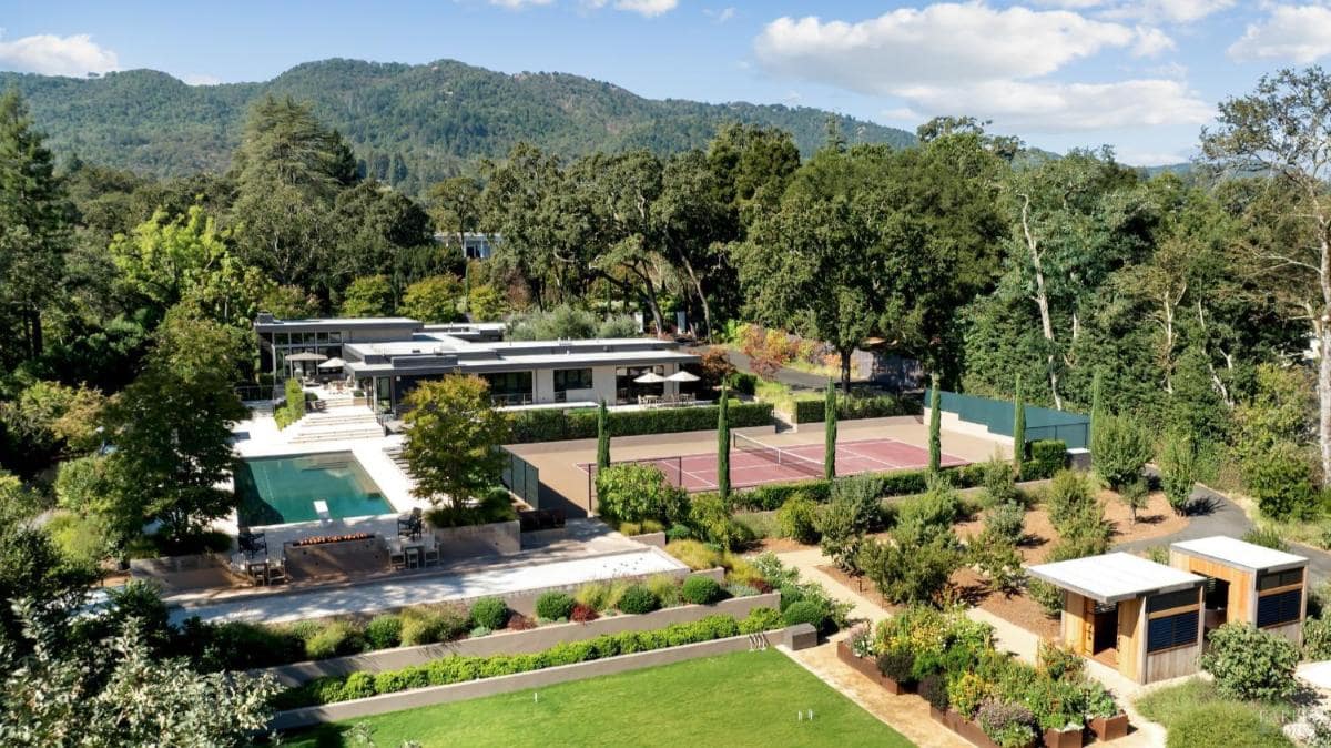 Aerial view of a property showing a swimming pool, tennis court, and landscaped garden with trees and hedges, set against a backdrop of hills.