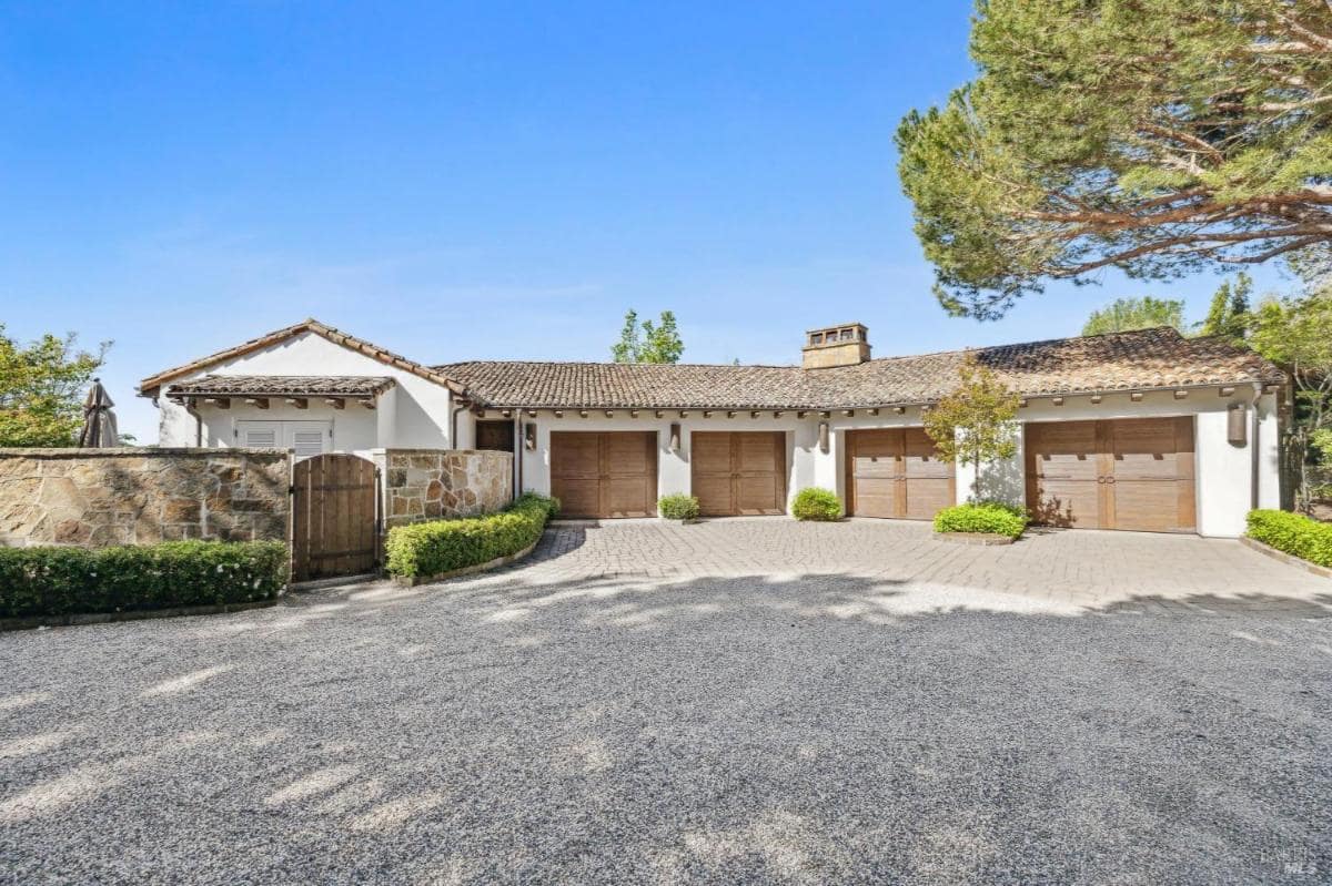 A garage with gravel driveway leads to the entrance, and there are landscaped areas with shrubs and trees.