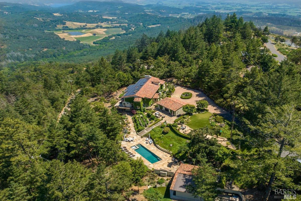 An aerial view of a house, pool, and surrounding forested area with landscaped grounds.