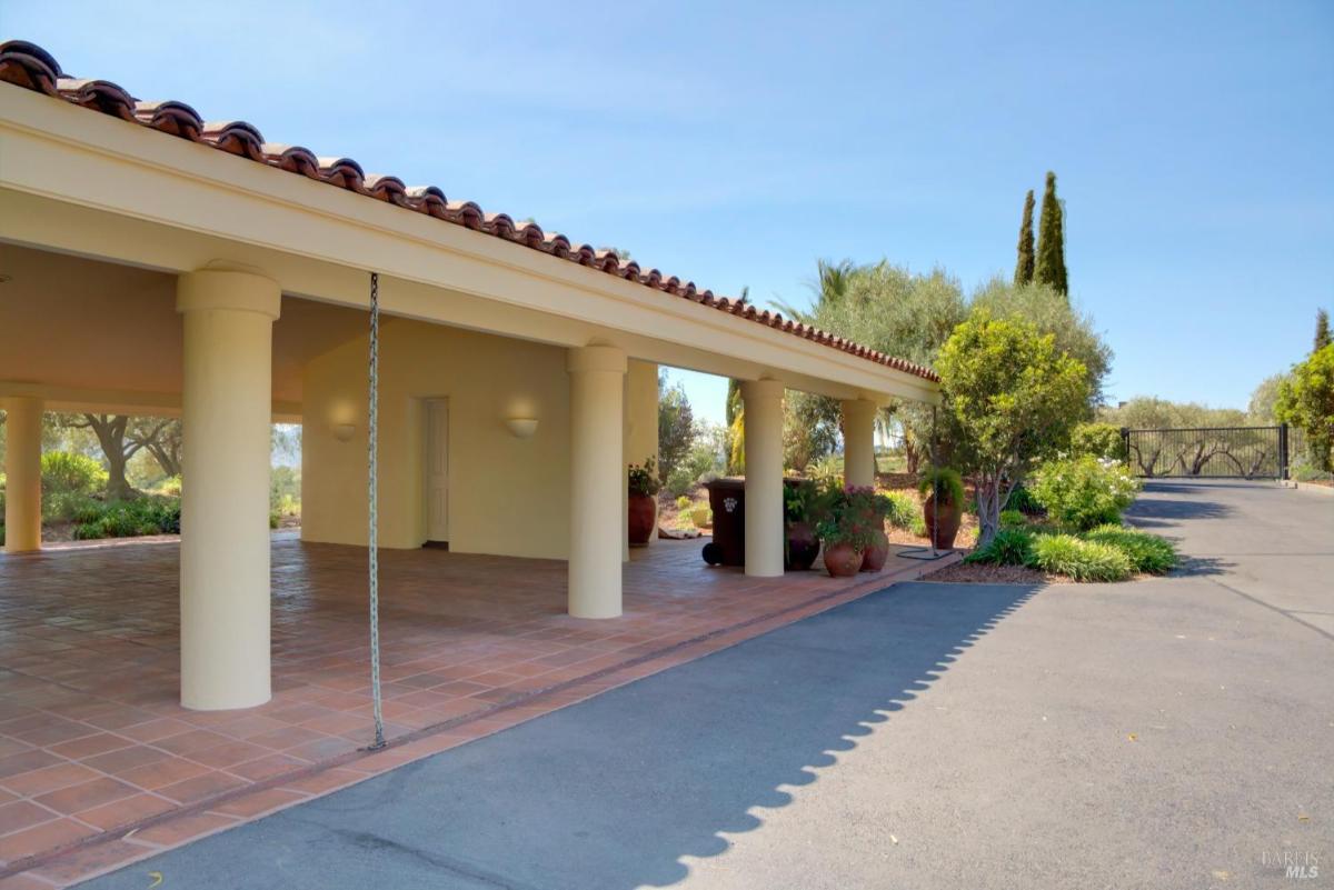 Covered open space with tiled flooring and supporting columns, near a gated driveway.