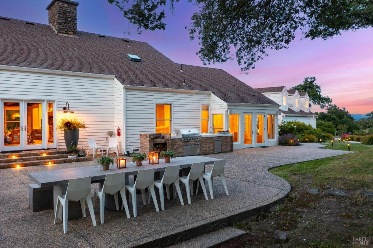 A house exterior includes a dining table, outdoor grill, and chairs, with evening lighting visible through the window